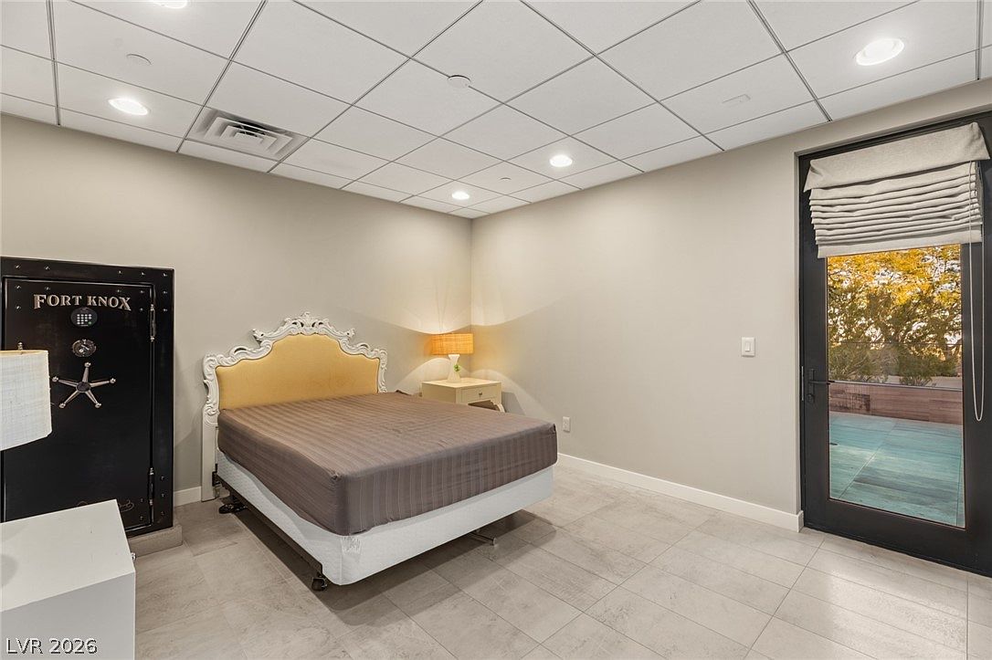 This bedroom features a neutral color palette with light-colored tile flooring and a drop ceiling. A bed with a distinctive, ornate white headboard and a yellow upholstered panel serves as the focal point, flanked by a small nightstand with a lamp. To the left, a large, black Fort Knox safe stands against the wall, while a glass door on the right provides access to an outdoor deck area.