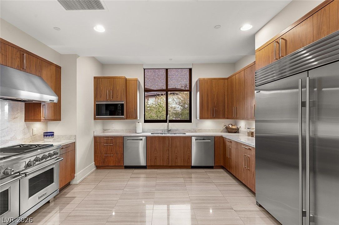 This spacious, U-shaped kitchen features warm-toned wood cabinetry and sleek stainless steel appliances, including a large professional-style range and a side-by-side refrigerator. A central window above the sink provides natural light, while recessed lighting illuminates the neutral stone countertops and polished tile flooring. The clean, contemporary design offers a functional layout with ample storage and workspace, creating an inviting atmosphere for cooking and entertaining.