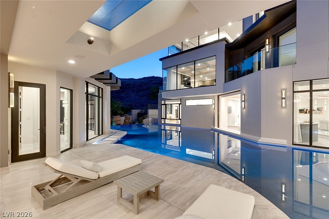 This luxurious outdoor living space features a sleek, modern infinity-edge pool that seamlessly integrates with the architecture of the home. A covered patio area with light-toned stone flooring provides a comfortable lounge space with chaise chairs, while large glass walls allow for an open connection between the interior and the pool deck. The scene is captured at dusk, highlighting the ambient lighting of the residence against the backdrop of a rugged hillside.
