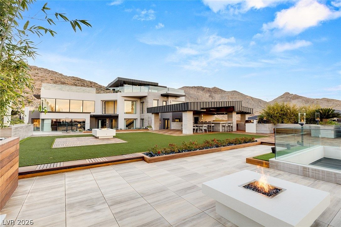 This expansive modern backyard features a large artificial turf lawn, a covered outdoor kitchen area with bar seating, and a sleek fire pit set into a tiled patio. The multi-level architectural design of the home includes large glass panels and a rooftop terrace, all set against a dramatic desert mountain backdrop. The scene is captured from a slightly elevated perspective, emphasizing the seamless transition between the indoor and outdoor living spaces.
