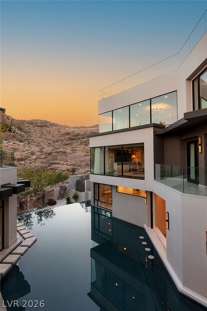 This striking modern residence features a multi-level architectural design with expansive glass walls that reflect the surrounding desert landscape. A sleek infinity-edge pool occupies the foreground, complete with integrated submerged seating, creating a seamless transition between the home and the rugged hillside backdrop. The composition captures a serene twilight atmosphere, highlighting the clean lines and sophisticated indoor-outdoor connectivity of the property.