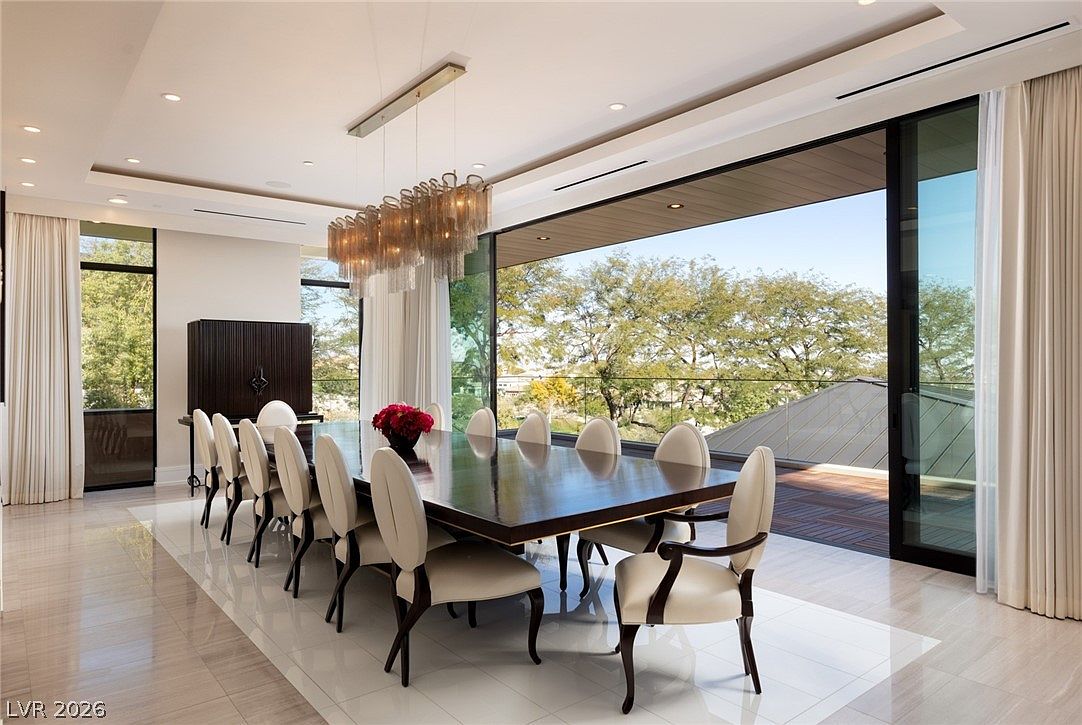 This elegant dining room features a long, dark-toned table surrounded by ten cream-colored upholstered chairs, centered beneath a striking tiered chandelier. The space is defined by large, floor-to-ceiling glass sliding doors that open onto an expansive outdoor deck, blurring the lines between indoor and outdoor living. The room is finished with light-colored tile flooring and neutral walls, creating a sophisticated and airy atmosphere perfect for formal entertaining.
