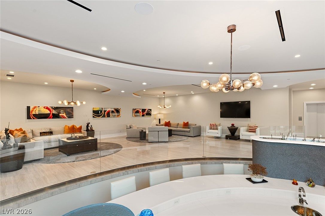 This expansive, open-concept living area features a sunken seating arrangement with plush white sofas and orange accent pillows, set against a backdrop of neutral walls and polished stone flooring. The space is illuminated by multiple modern, multi-globe chandeliers and recessed lighting, creating a sophisticated and airy atmosphere. A glass railing separates the lower seating area from the foreground, where a curved white counter suggests a bar or kitchen island, emphasizing the home's spacious and contemporary design.