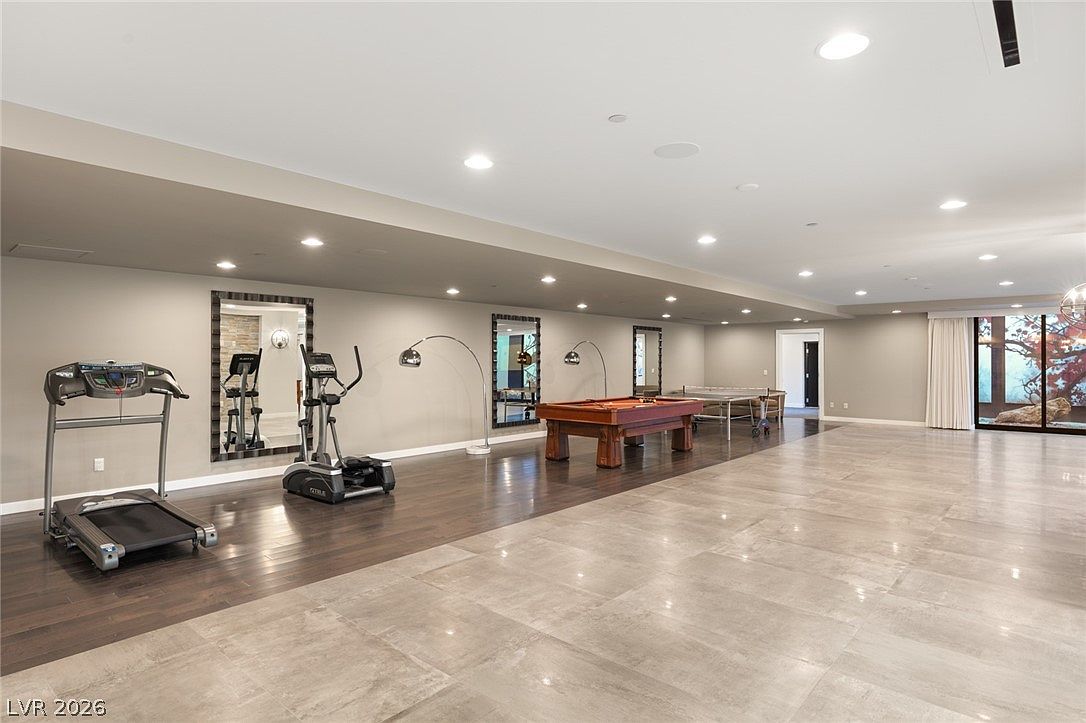 This expansive, multi-purpose recreation room features a combination of dark wood flooring and large, light-toned tiles. The space is equipped with a treadmill and elliptical machine on one side, while a pool table and ping-pong table occupy the center, creating a versatile area for fitness and entertainment. Recessed lighting illuminates the room, which also includes large mirrors and a sliding glass door that provides a view of the outdoor landscape.