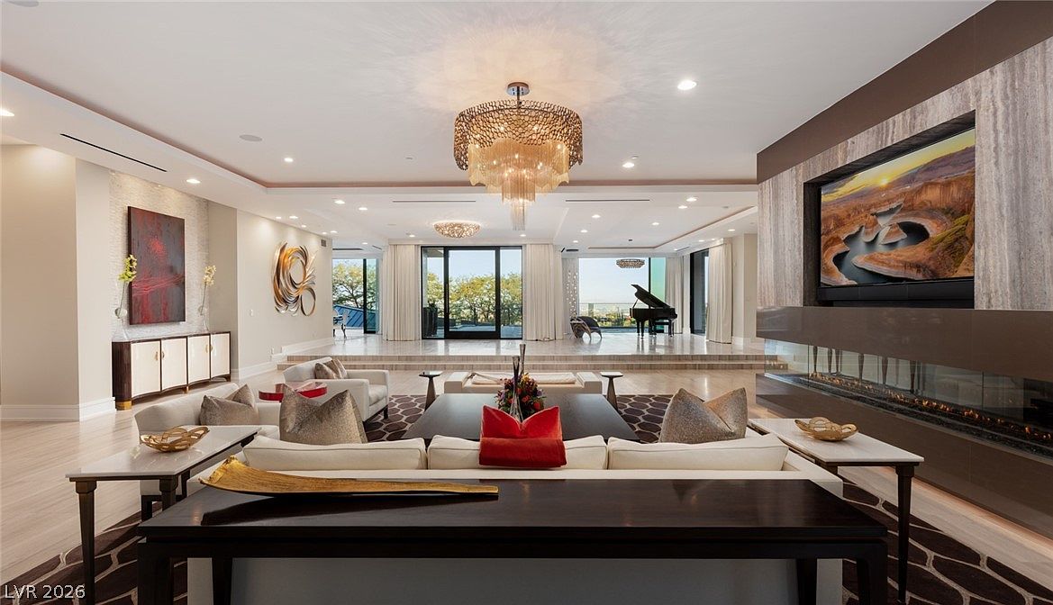 This expansive, open-concept living room features a sophisticated design with a sunken seating area and a raised platform leading to a grand piano. A prominent stone-clad chimney breast houses a wide, linear fireplace and a large television, while a tiered crystal chandelier serves as a dramatic focal point. The space is framed by large windows that offer a seamless transition to the outdoor scenery, creating an atmosphere of luxury and refined elegance.