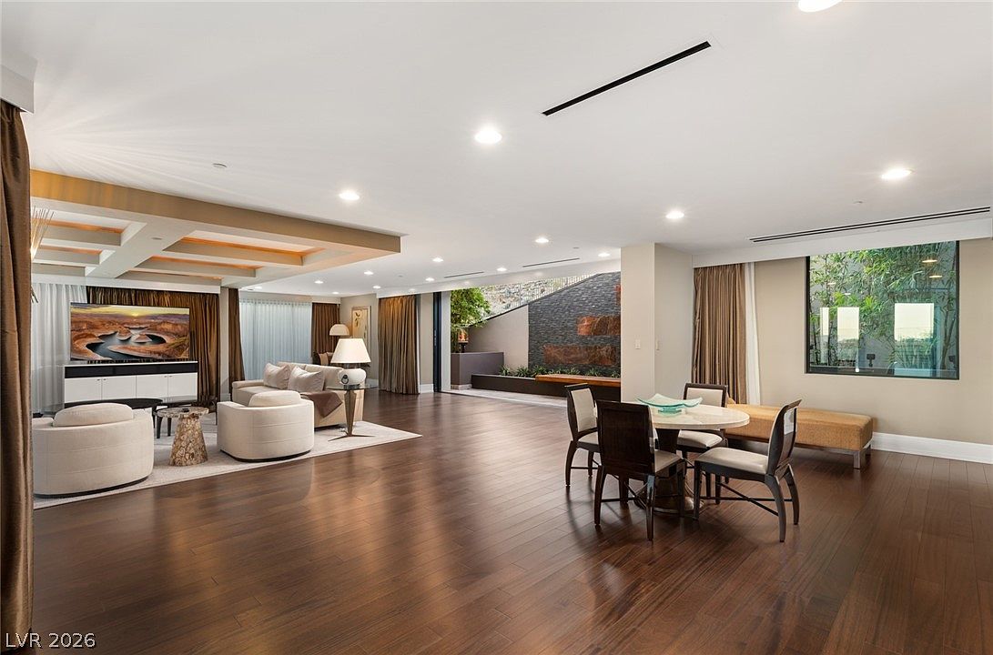 This expansive open-concept living and dining area features rich dark wood flooring and a neutral color palette. The living space is anchored by a large television console and comfortable seating, while the adjacent dining area includes a circular table and chairs. A large glass opening leads to an outdoor patio with a stone-accented water feature, creating a seamless transition between indoor and outdoor living spaces.