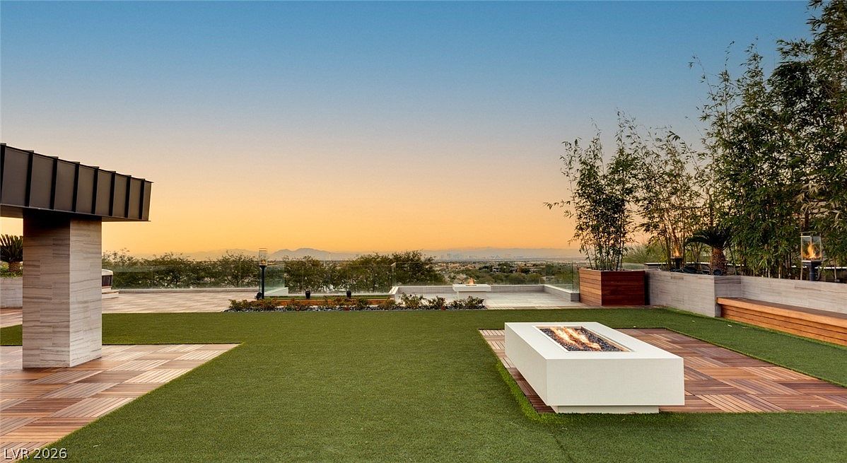 This expansive rooftop terrace features a modern fire pit as a central focal point, set against a backdrop of panoramic mountain and city views. The space is finished with a combination of artificial turf and wood-plank decking, complemented by tall bamboo planters and a sleek glass railing. The scene is captured at sunset, creating a warm, inviting atmosphere perfect for outdoor entertaining.