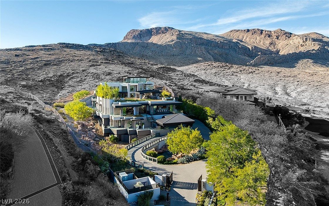 This high-angle aerial view captures a sprawling, multi-level contemporary estate nestled against a rugged, arid mountain backdrop. The architecture features clean lines, expansive terraces, and a sophisticated terraced design that integrates seamlessly with the desert landscape. A winding paved driveway leads to the residence, surrounded by manicured desert landscaping and mature trees, creating a striking contrast between the modern structure and the natural terrain.
