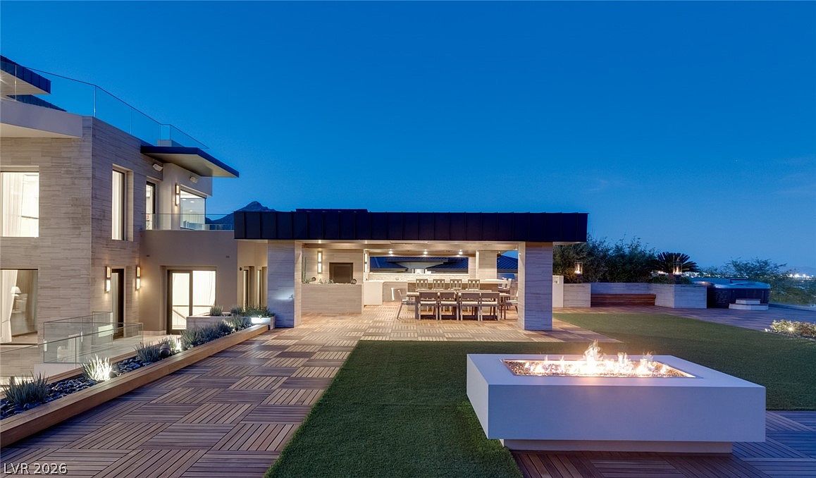 This expansive outdoor living space features a modern covered patio area with a dining set, seamlessly transitioning to a deck with a contemporary rectangular fire pit. The architecture showcases clean lines, light-toned stone walls, and large glass panels, creating a sophisticated and airy atmosphere. The scene is captured at dusk, highlighting the warm glow of the fire pit and recessed lighting against the deep blue twilight sky.