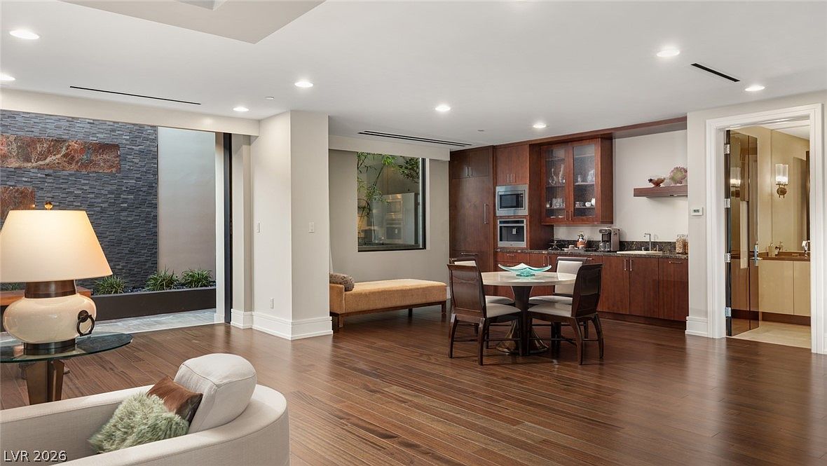 This spacious open-concept living area features rich, dark wood flooring and a neutral color palette that creates an inviting atmosphere. The room includes a comfortable seating area with a curved sofa, a small dining set near a wet bar with dark cabinetry, and a unique indoor feature wall with textured stone cladding. Recessed lighting throughout the ceiling provides a bright, modern feel, while the layout flows seamlessly into an adjacent room through a doorway.