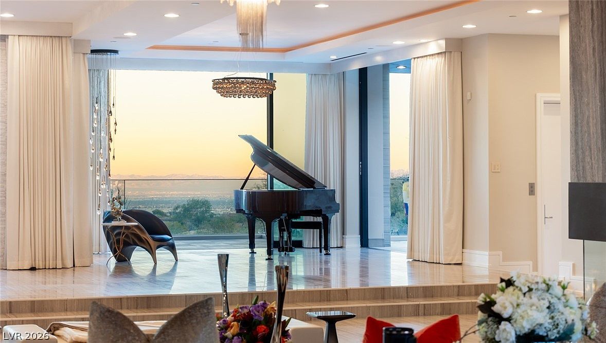 This sophisticated living space features a raised platform area showcasing a grand piano positioned against expansive glass walls that offer a panoramic view of the distant mountains. The room is characterized by a neutral color palette, elegant drapery, and a tiered layout that creates a sense of depth and luxury. A modern chandelier hangs above the piano, while the foreground includes plush seating and floral arrangements, emphasizing a high-end, cinematic atmosphere.
