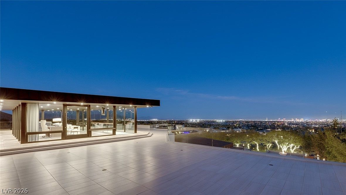This expansive, modern terrace features large-format light-colored tiles and a sleek glass railing, offering an unobstructed panoramic view of the city lights at dusk. A contemporary structure with floor-to-ceiling glass walls opens directly onto the deck, creating a seamless transition between indoor and outdoor living spaces. The minimalist design and elevated vantage point emphasize a luxurious, sophisticated atmosphere.