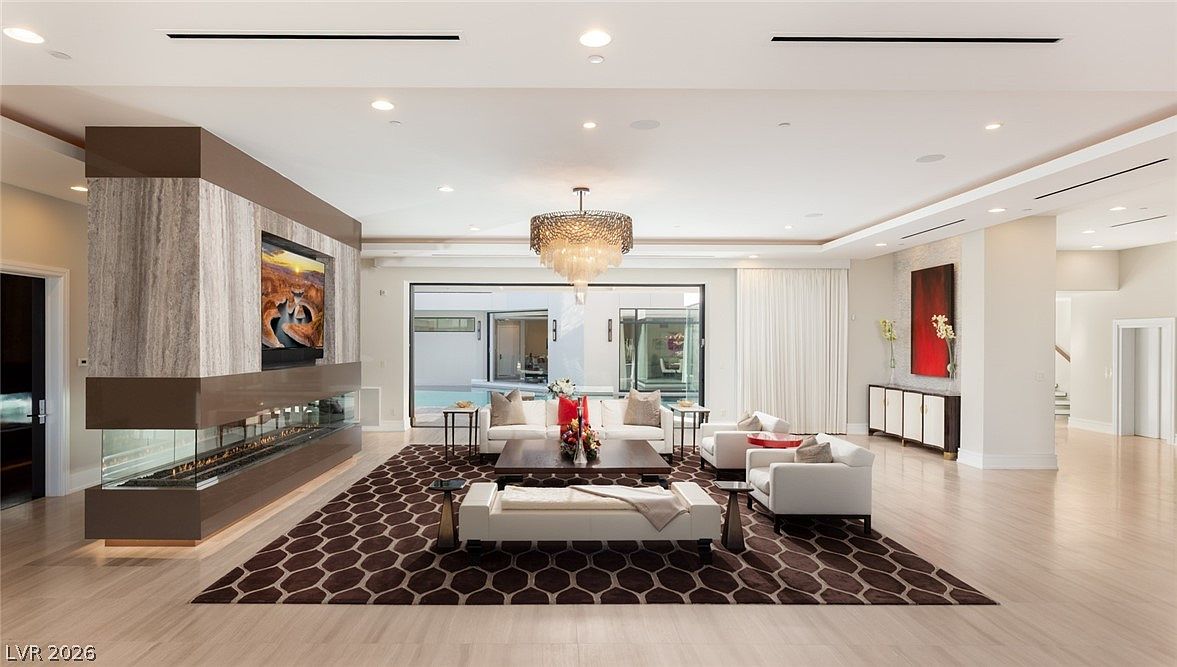 This expansive, open-concept living room features a sophisticated neutral color palette and a large geometric-patterned area rug that anchors the seating area. A prominent stone-clad chimney breast with a built-in television and a linear fireplace serves as a central architectural feature, while large glass doors provide a seamless transition to the outdoor pool area. The space is illuminated by a grand, multi-tiered crystal chandelier and recessed lighting, creating an atmosphere of refined luxury.
