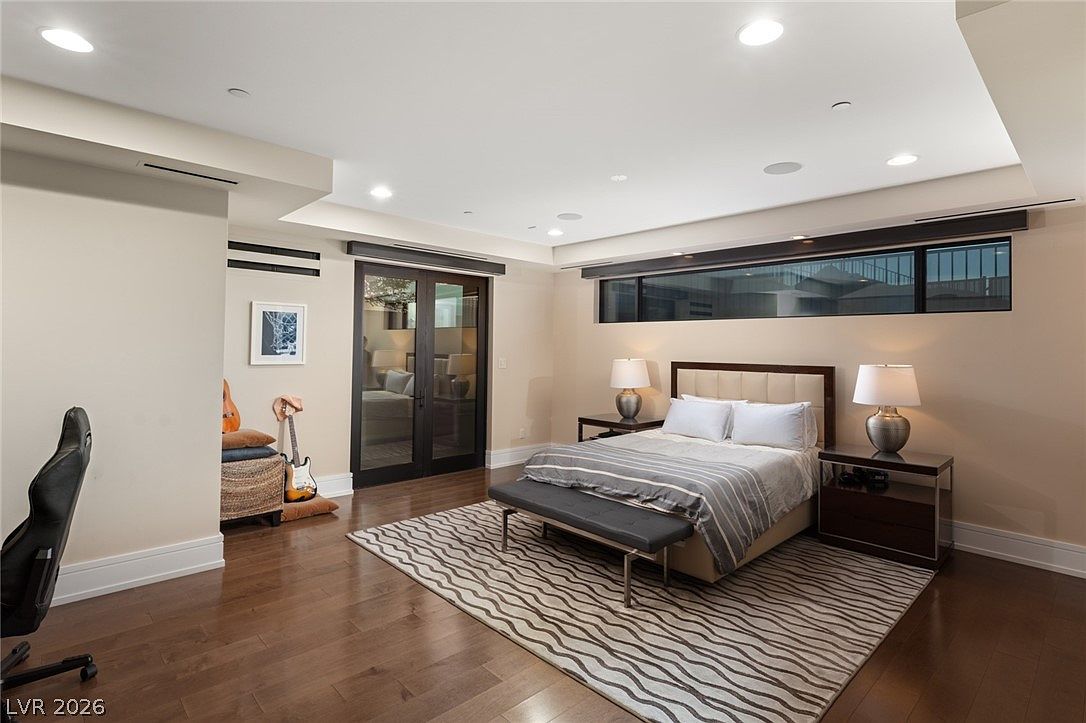 This spacious bedroom features dark hardwood flooring and a neutral color palette, creating a serene and modern atmosphere. The room is anchored by a bed with a tufted headboard, flanked by two nightstands with matching lamps, and includes a bench at the foot of the bed. A large area rug with a geometric pattern sits beneath the bed, while a set of French doors and a long, horizontal window provide natural light and a view into an adjacent space.