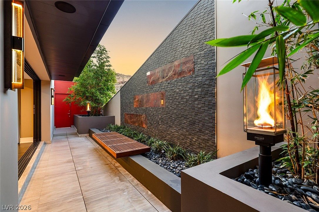 This modern outdoor patio space features a sleek, linear design with large-format light-toned tiles and a striking dark stone-clad feature wall accented by rectangular stone inlays. A built-in planter bed with lush greenery and a contemporary glass-enclosed fire feature creates a sophisticated atmosphere, while a wooden bench provides integrated seating. The perspective looks down the length of the covered walkway toward a vibrant red door, emphasizing the clean lines and high-end architectural aesthetic.