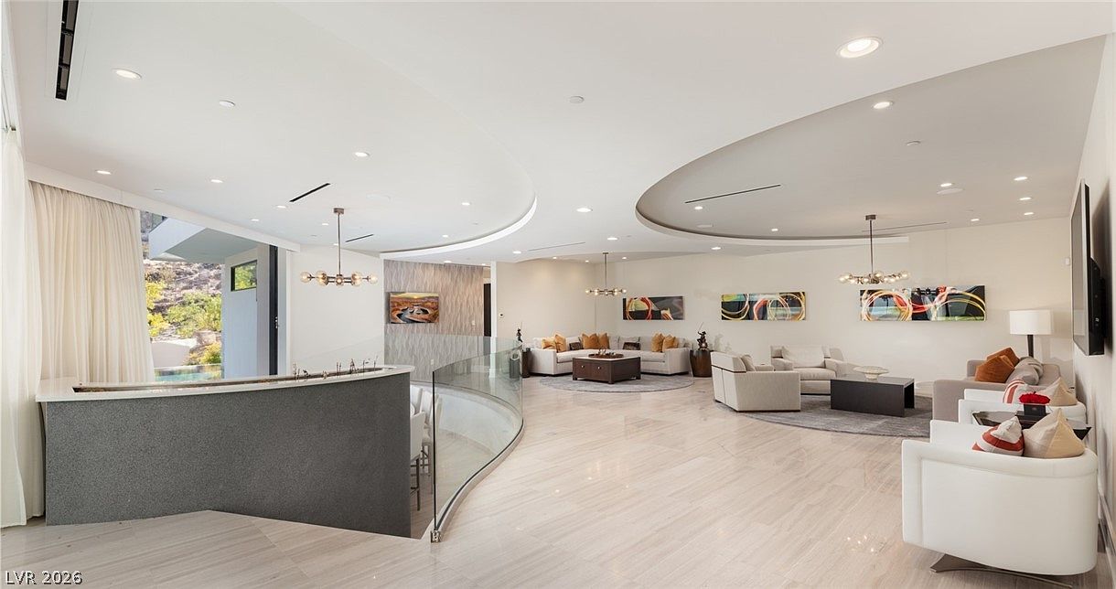 This expansive, open-concept living area features a sophisticated, modern design with a curved architectural ceiling detail and polished light-toned flooring. The space is furnished with multiple seating arrangements, including plush sofas and armchairs, and is decorated with vibrant abstract artwork. Large glass panels provide a seamless transition to the outdoor area, creating a bright and airy atmosphere that emphasizes luxury and spaciousness.