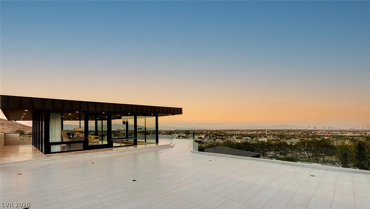 This expansive, modern terrace features light-toned tiled flooring and a sleek, minimalist structure with floor-to-ceiling glass walls that offer an unobstructed view of the surrounding landscape and distant city skyline. The architectural design emphasizes clean lines and a seamless transition between the indoor office space and the outdoor living area. The scene is captured during a serene sunset, casting a warm, golden glow over the entire property.