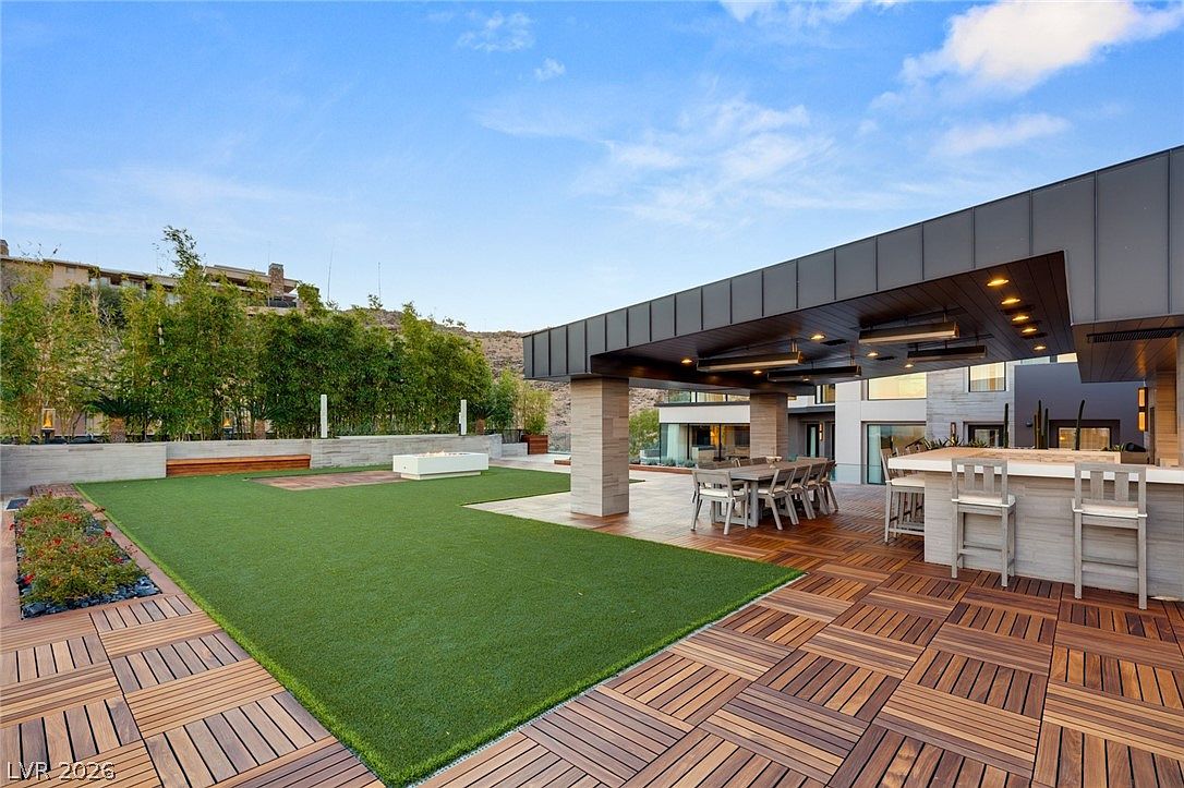 This expansive outdoor living space features a modern covered patio with a built-in bar and dining area, seamlessly transitioning to a manicured artificial turf lawn. The deck is constructed with modular wood tiles, and a minimalist fire feature sits centrally on the lawn, framed by lush bamboo landscaping. The perspective captures a sophisticated, resort-style entertainment area designed for seamless indoor-outdoor living.
