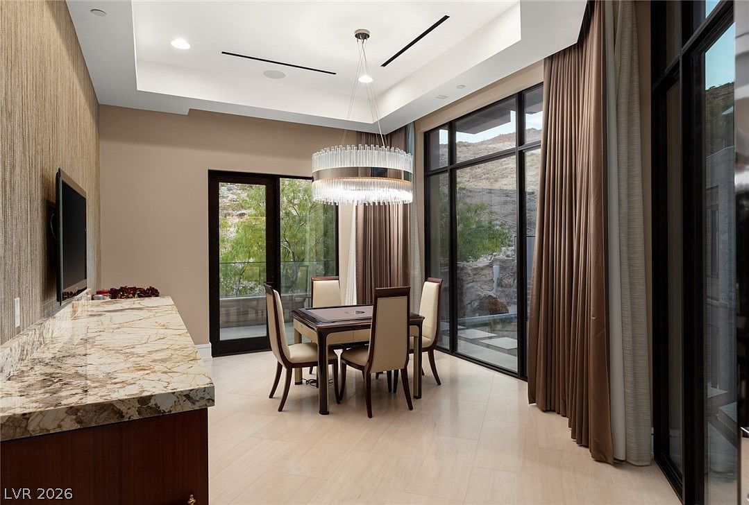 This elegant dining area features a square table surrounded by four upholstered chairs, positioned beneath a modern, tiered crystal chandelier. The room is framed by expansive glass walls that offer views of the rugged, rocky landscape outside, complemented by floor-to-ceiling curtains. To the left, a long stone-topped console provides additional surface space, while the neutral color palette and recessed tray ceiling create a sophisticated and airy atmosphere.