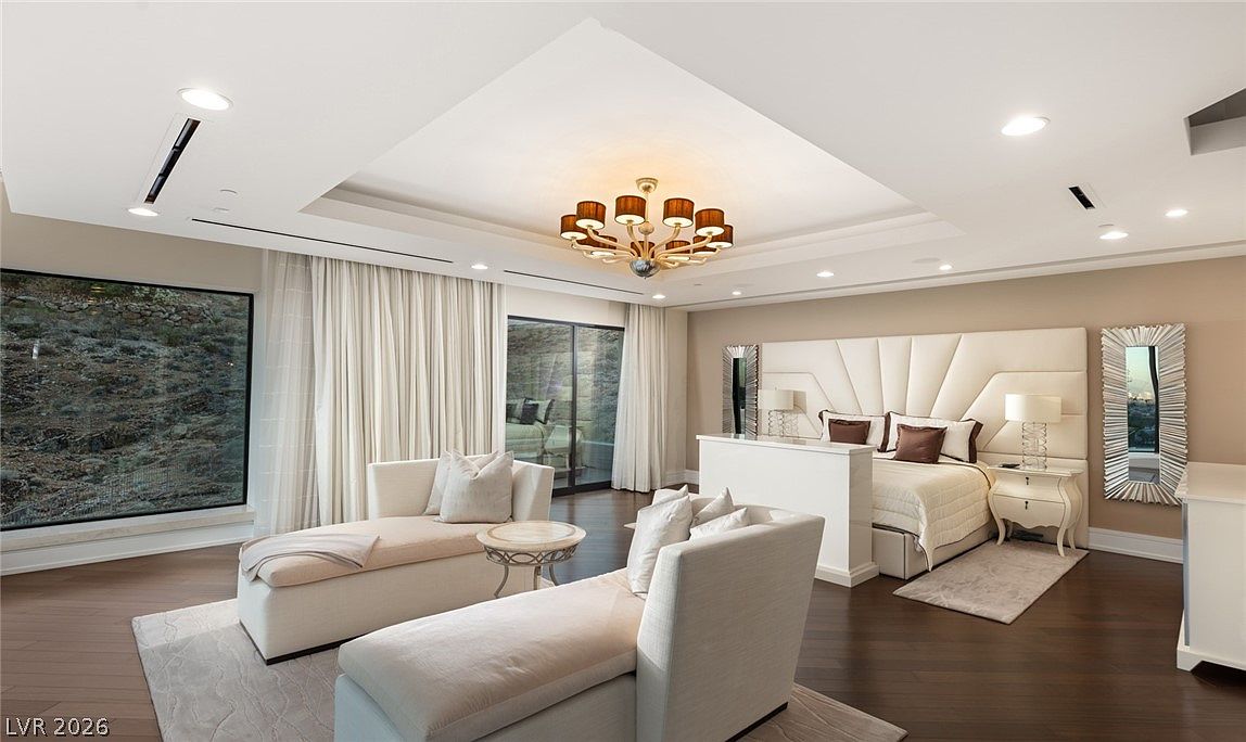 This expansive primary bedroom features a sophisticated neutral palette with a large, tufted upholstered headboard as the focal point. The room includes a dedicated seating area with two chaise lounges, dark wood flooring, and a decorative chandelier suspended from a recessed ceiling tray. Large windows provide natural light and views of the exterior landscape, creating an airy and luxurious atmosphere.