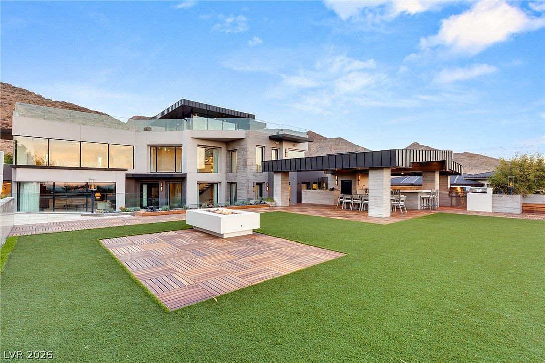 This modern luxury residence features a multi-level architectural design with expansive glass walls and a sleek, contemporary aesthetic. The backyard includes a manicured lawn, a central fire pit area with a wood-plank deck, and a covered outdoor kitchen and dining space. The property is set against a dramatic mountain backdrop, emphasizing a seamless transition between indoor and outdoor living.