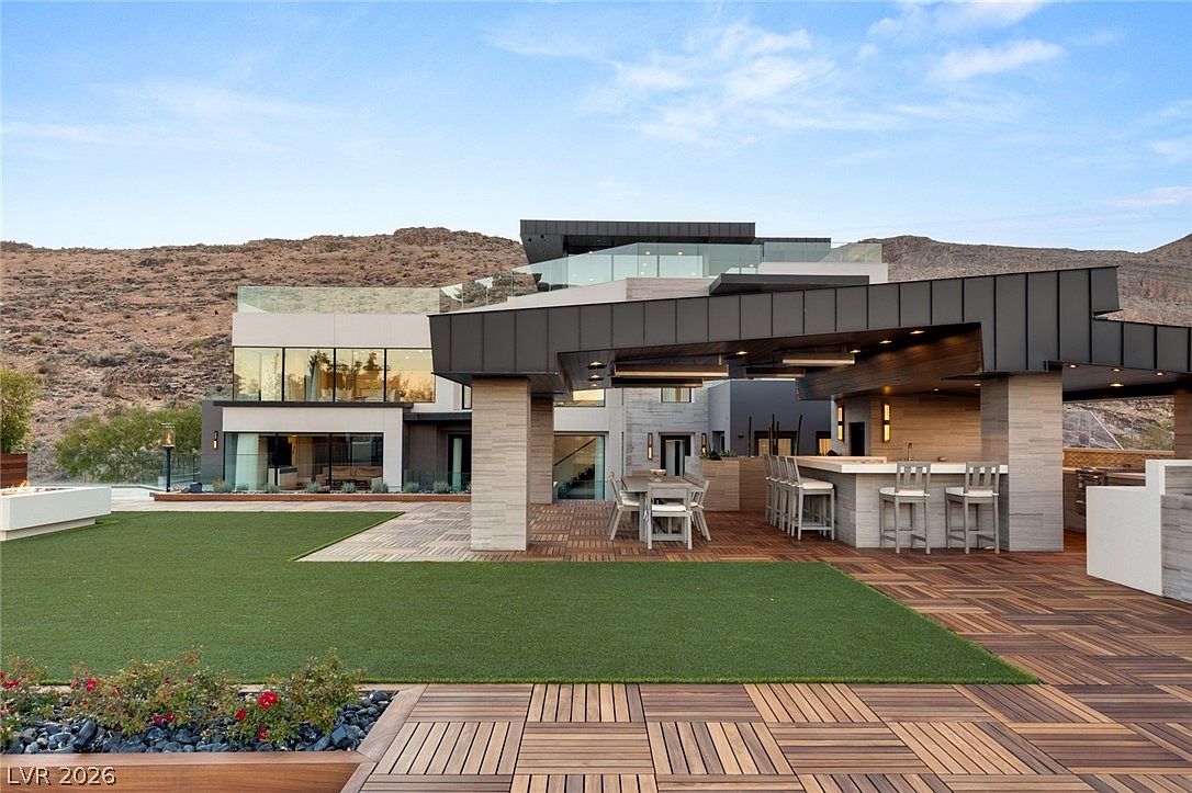 This expansive outdoor living space features a modern covered patio with a built-in bar and dining area, set against a backdrop of rugged desert mountains. The area includes a large section of artificial turf, wood-tiled decking, and a contemporary multi-level home with large windows and glass railings. The scene is captured from a low-angle perspective, emphasizing the seamless transition between the indoor and outdoor entertainment zones.