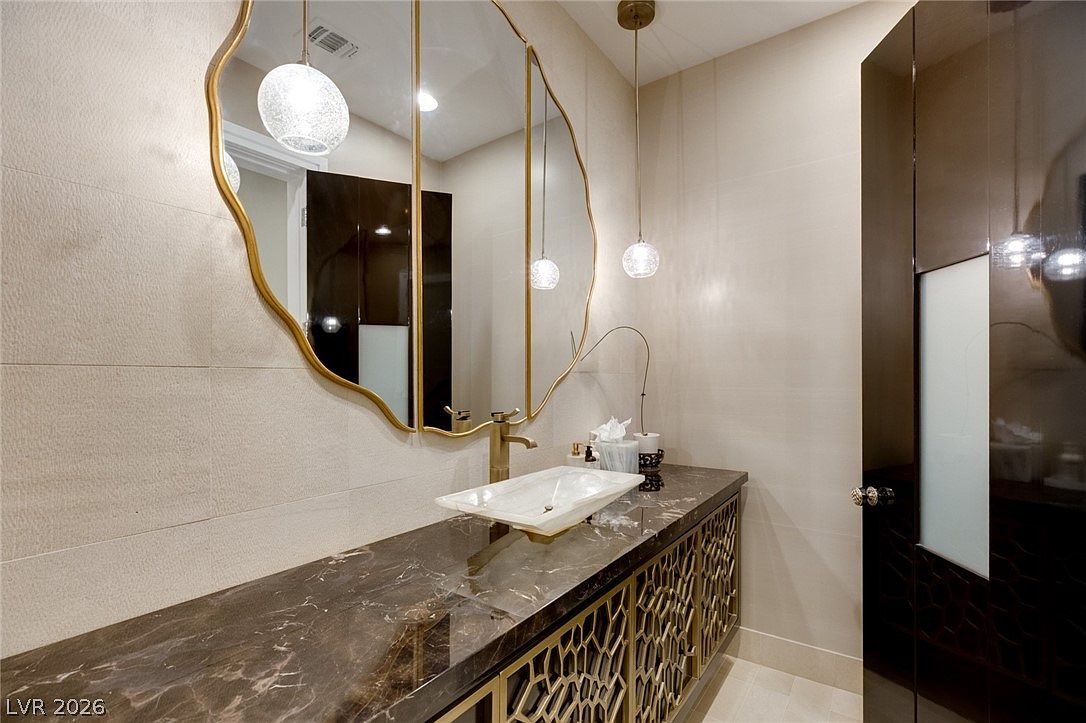 This elegant powder room features a dark stone vanity top with a unique, translucent vessel sink and a gold-toned faucet. A large, organic-shaped mirror with a gold frame hangs above the vanity, complemented by two modern pendant lights. The space is finished with light-colored wall tiles and a decorative gold-toned metal cabinet base, creating a sophisticated and luxurious atmosphere.