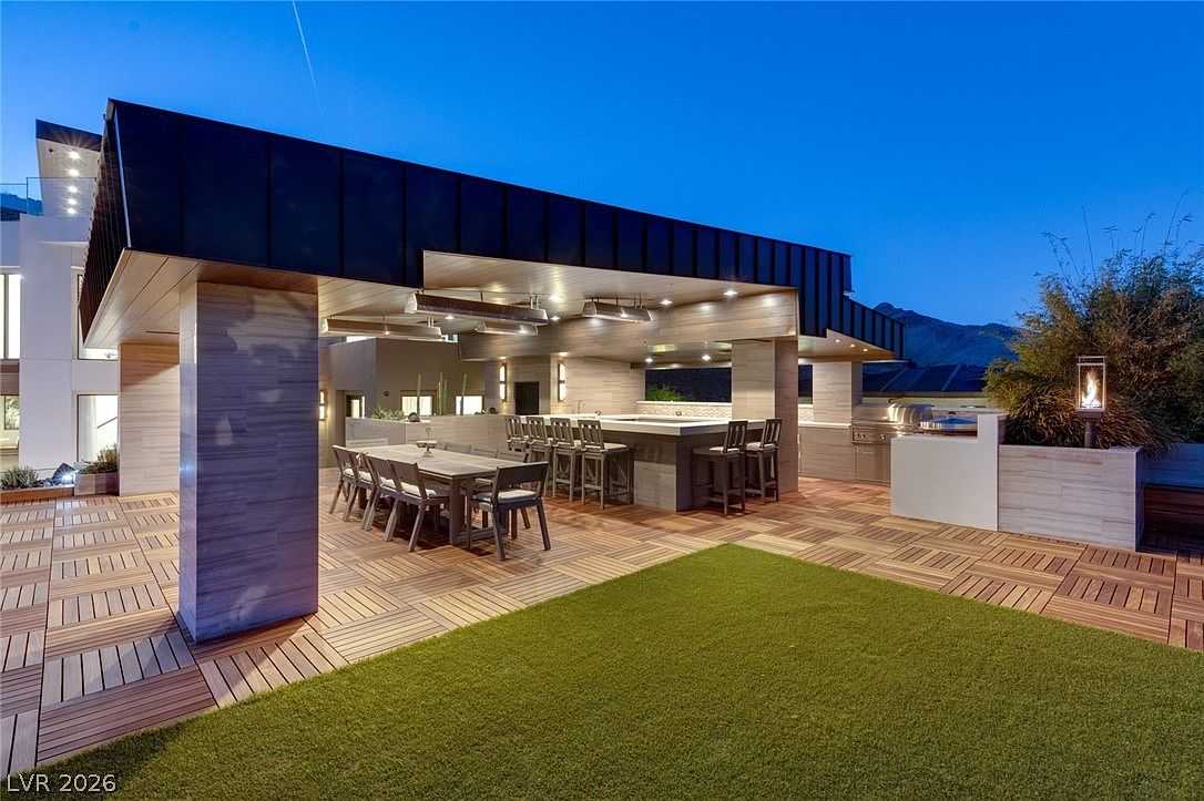 This expansive outdoor living space features a covered patio area with a large dining table and a built-in bar with barstool seating. The area is finished with patterned wood decking and overlooks a patch of manicured artificial turf, creating a seamless transition between indoor and outdoor entertaining zones. The modern architectural design includes clean lines, recessed lighting, and a sleek, dark-toned roof structure that provides shade for the kitchen and dining setup.