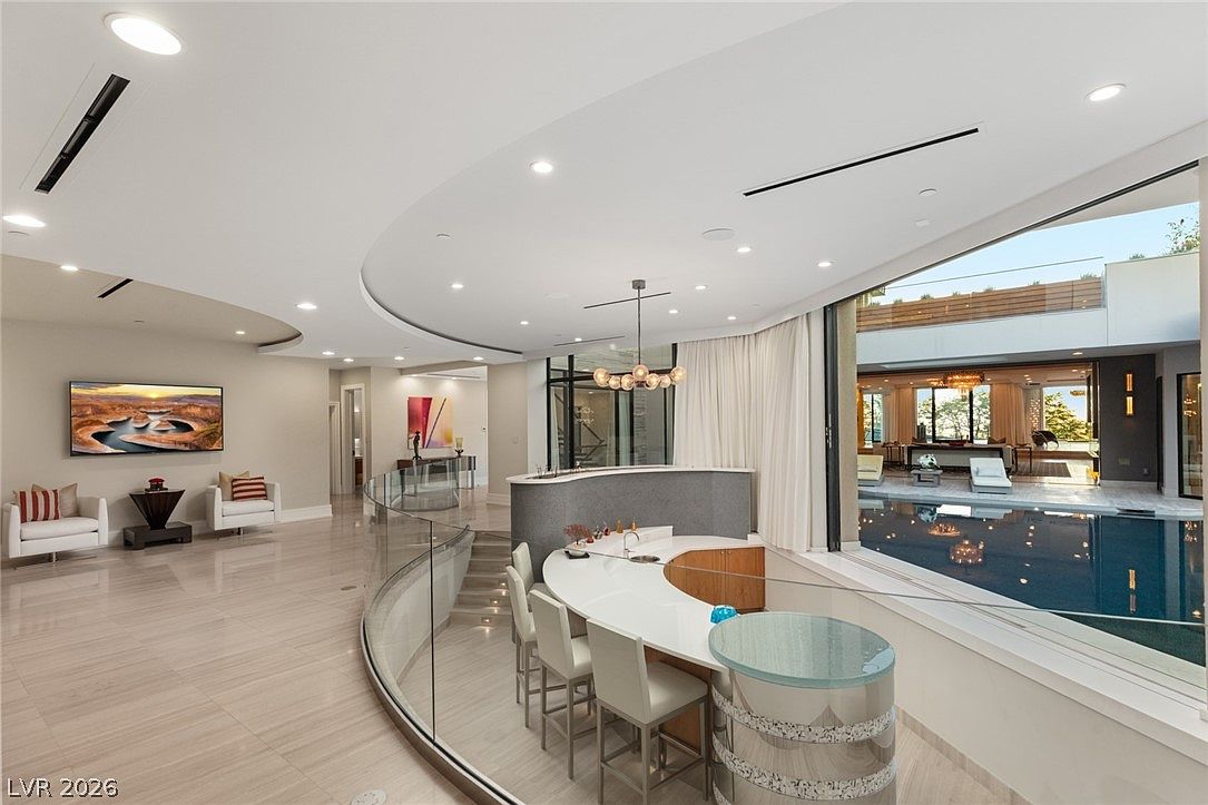 This expansive, modern living area features a curved, glass-railed staircase leading to a lower level and a central bar area with white countertops and bar seating. Large, sliding glass walls open directly onto an indoor-outdoor pool, creating a seamless transition between the interior living space and the water. The room is decorated in a neutral palette with contemporary furniture, recessed lighting, and a large landscape artwork on the wall.
