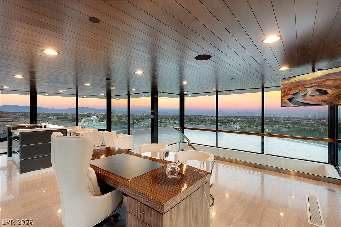 This sophisticated office space features a polished wood desk and modern white seating, set against a backdrop of expansive floor-to-ceiling windows that offer a panoramic view of the city and mountains at sunset. The room is defined by a dark, slatted wood ceiling and glossy light-toned flooring, creating a sleek, high-end atmosphere. A glass railing separates the office area from an adjacent outdoor terrace, emphasizing the seamless transition between indoor and outdoor living.