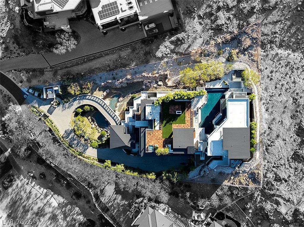 This high-angle aerial view captures a sprawling, modern luxury estate nestled into a rugged desert landscape. The property features a complex architectural footprint with multiple flat-roofed sections, a large swimming pool, a central courtyard with green space, and a curved driveway leading to a private entrance. The surrounding terrain is rocky and arid, emphasizing the home's secluded and elevated position.