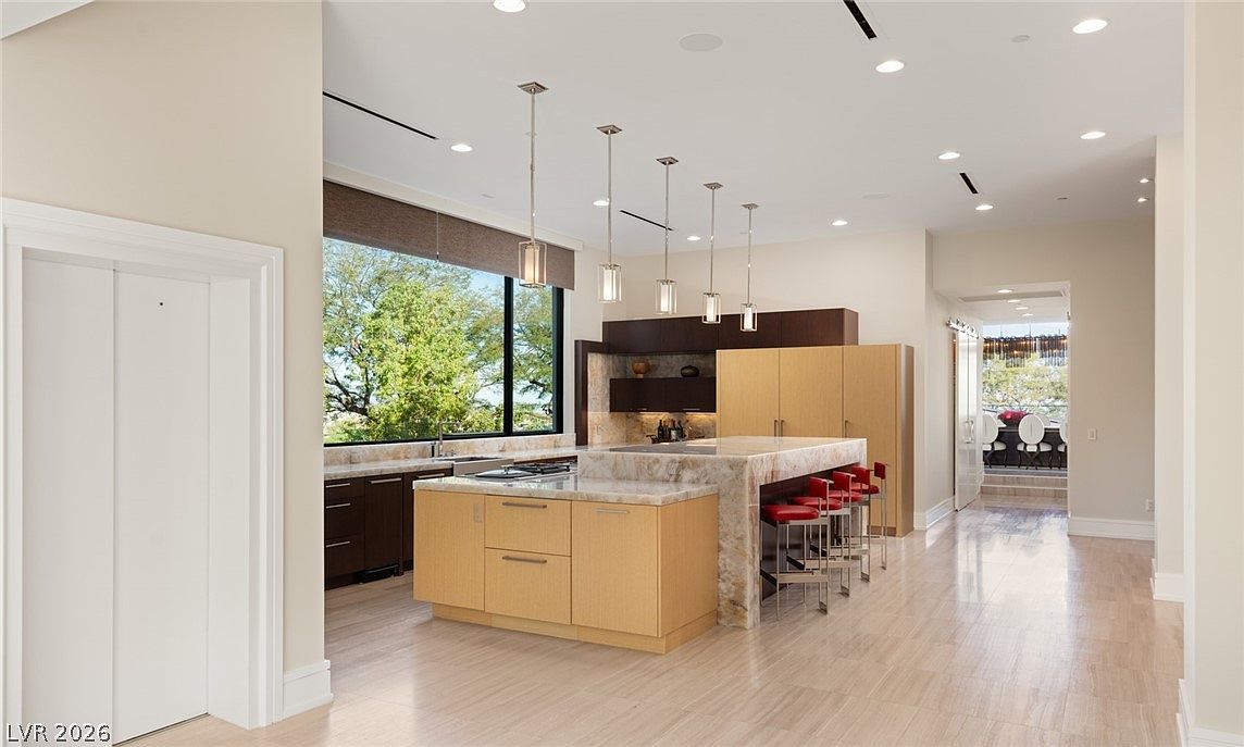 This spacious, modern kitchen features a large central island with a stone countertop and light wood cabinetry, complemented by a row of red bar stools. The room is illuminated by a series of pendant lights hanging above the island and recessed ceiling lighting, creating a bright and open atmosphere. A large window provides a view of the outdoors, while a doorway in the background leads to an adjacent balcony area.