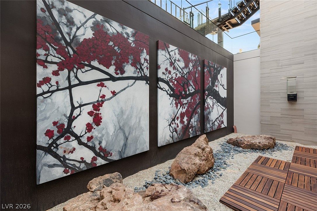 This serene outdoor courtyard features a striking triptych of cherry blossom-themed artwork mounted on a dark, modern wall. The ground is composed of light-colored gravel accented by large, natural boulders and a section of wooden decking, creating a minimalist Zen-inspired aesthetic. A glass-railed staircase is visible in the upper background, adding a contemporary architectural element to the tranquil space.