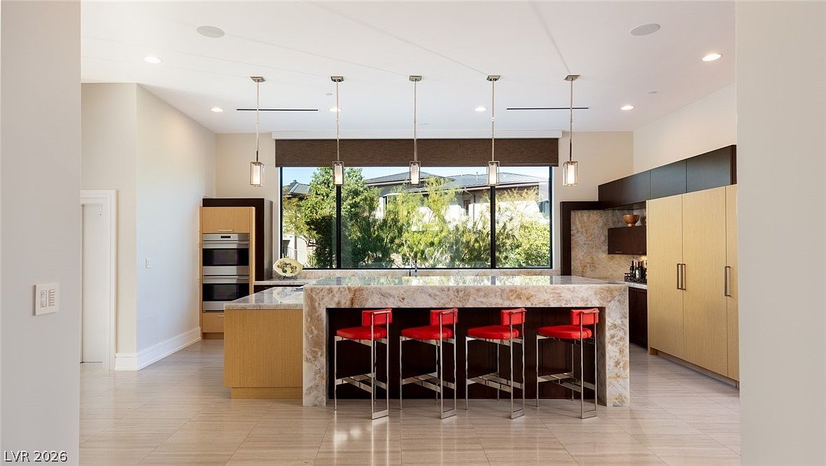 This modern kitchen features a large central island with a stone countertop and four vibrant red bar stools, serving as the focal point of the room. The space is illuminated by four sleek pendant lights hanging above the island and recessed ceiling lighting, while a large window provides a view of the outdoors. The kitchen is equipped with built-in appliances and a mix of light wood and dark cabinetry, creating a clean, contemporary aesthetic.