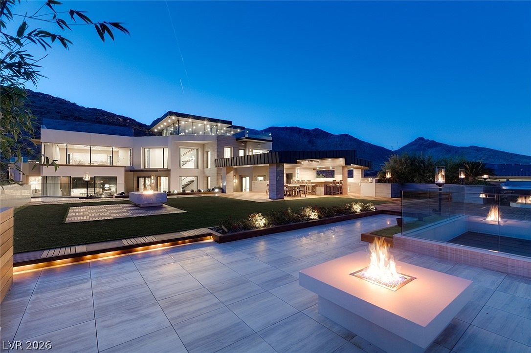 This stunning contemporary estate is captured at twilight, showcasing a sprawling rear patio with multiple fire features and a manicured lawn. The architecture features clean lines, expansive glass walls, and a covered outdoor dining area, all set against a dramatic mountain backdrop. The scene is illuminated by warm landscape lighting, creating an inviting and luxurious atmosphere for evening entertaining.