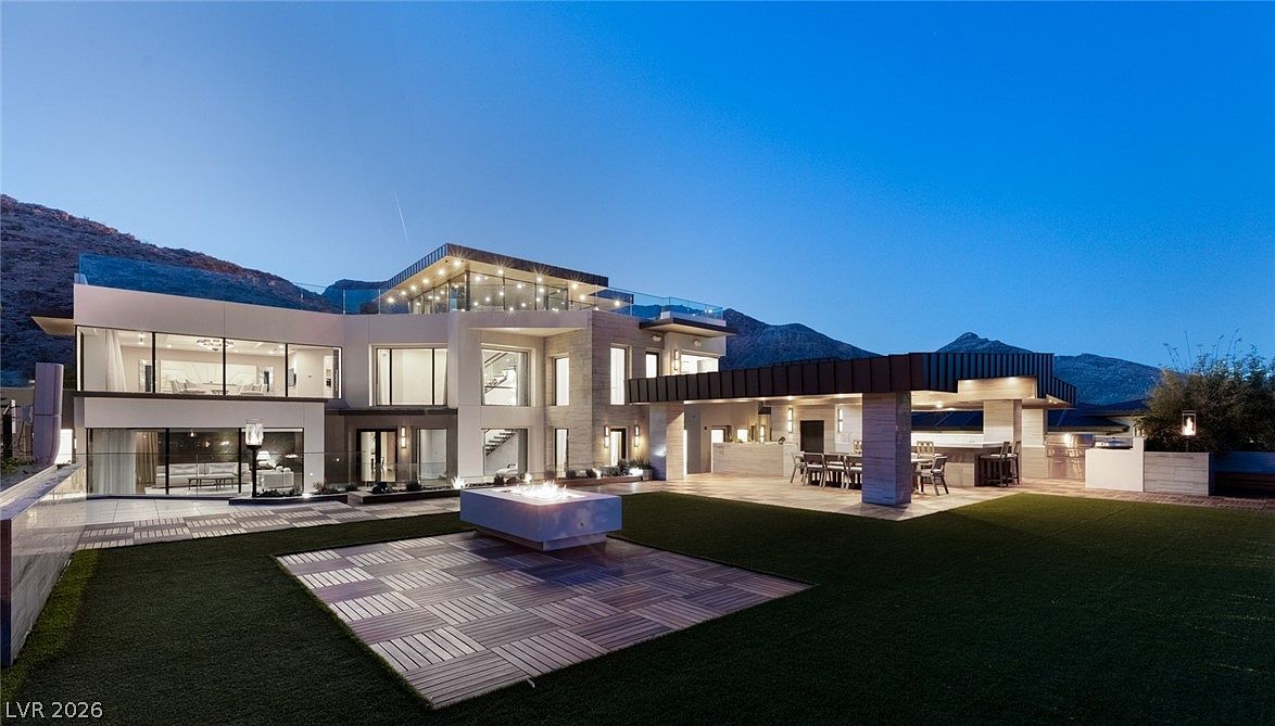 This modern luxury residence is captured from the rear yard at twilight, showcasing a multi-level architectural design with expansive glass walls and a rooftop terrace. The outdoor living space features a covered patio with a built-in bar, a central fire pit area, and a manicured lawn set against a dramatic mountain backdrop. The home's contemporary aesthetic is emphasized by clean lines, integrated lighting, and a seamless transition between indoor and outdoor living areas.