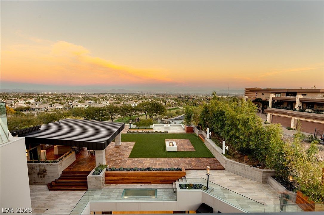 This elevated outdoor living space offers a panoramic view of the surrounding desert landscape and city skyline at sunset. The area features a covered patio with a dark metal roof, a central patch of artificial turf, and tiered stone-tiled decking. A glass railing provides an unobstructed view, while integrated lighting and modern landscaping elements create a sophisticated, high-end atmosphere.