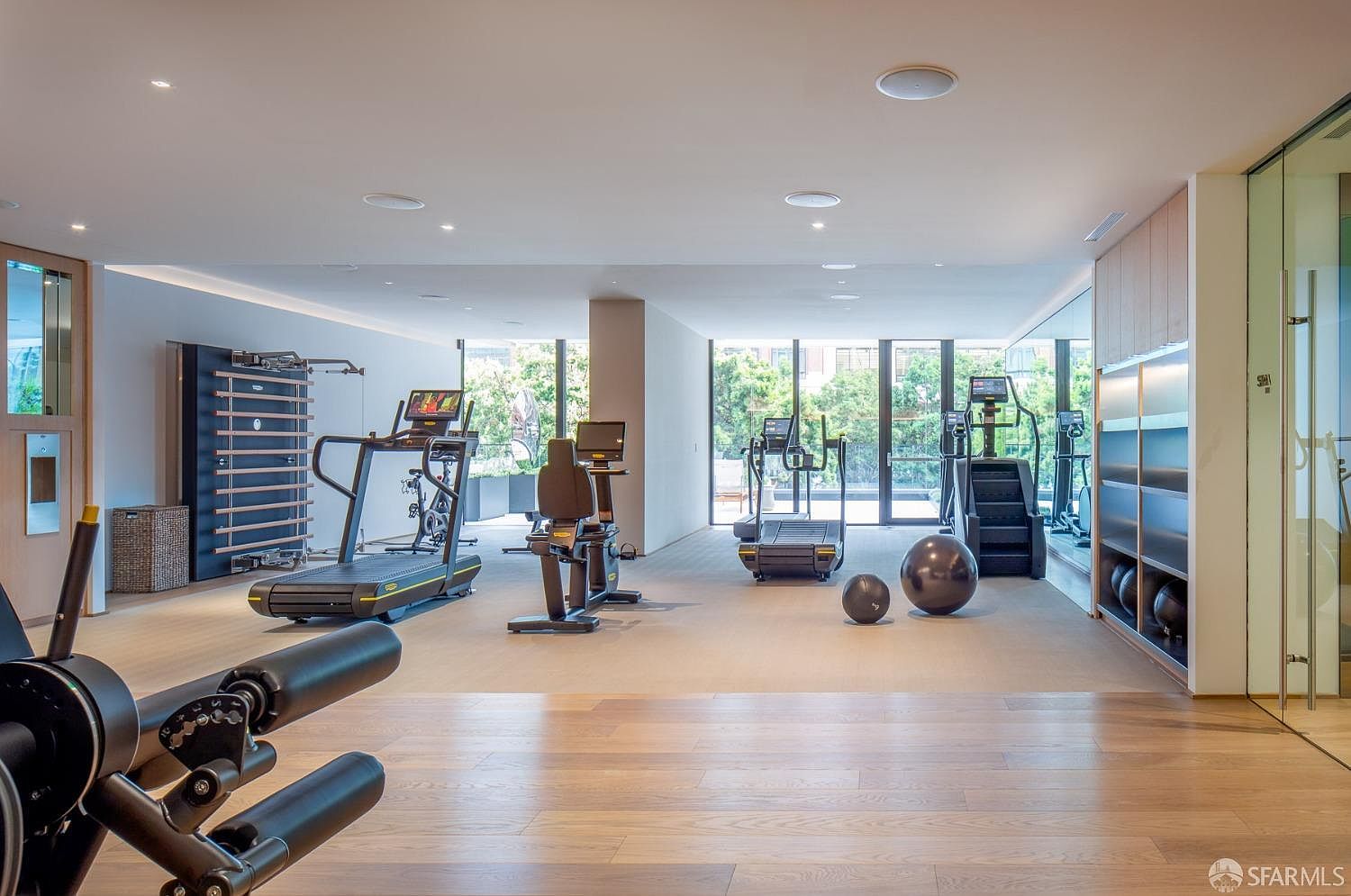 This is a well-equipped home gym featuring various exercise machines such as treadmills, stationary bikes, and a stair climber. The room has hardwood flooring, large windows providing natural light, and a mirrored wall. The overall impression is a clean, modern, and functional space for fitness activities.