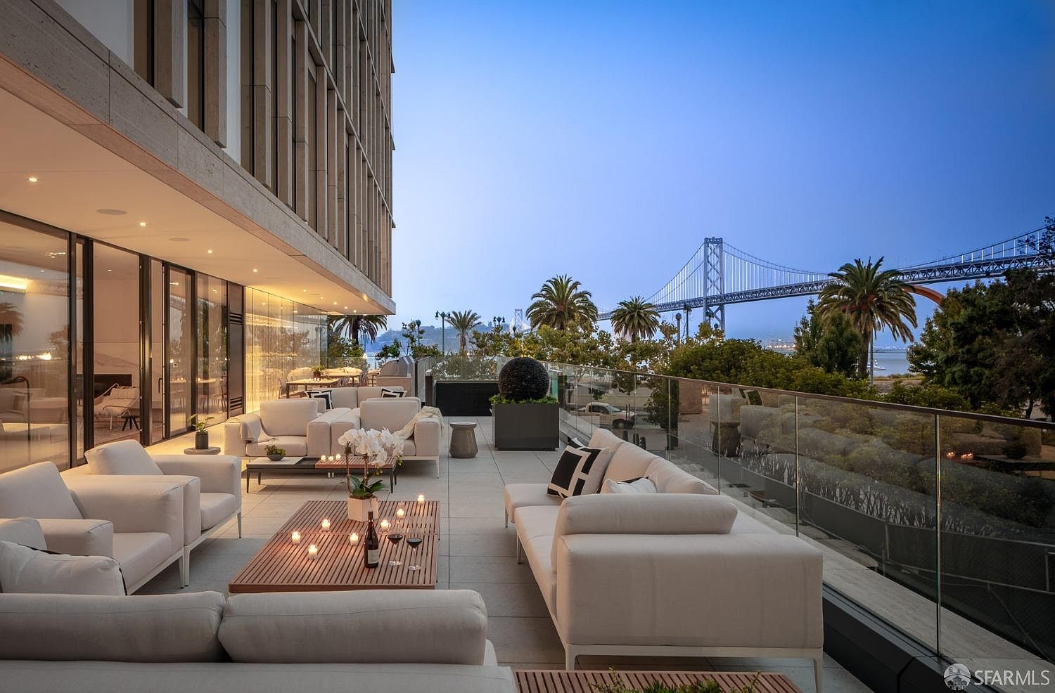 This image showcases a luxurious outdoor patio or balcony area, featuring modern white sofas and lounge furniture arranged around wooden coffee tables. The space is illuminated by ambient lighting, creating a warm and inviting atmosphere. A glass railing provides unobstructed views of the city skyline and a distant bridge, enhancing the property's appeal.