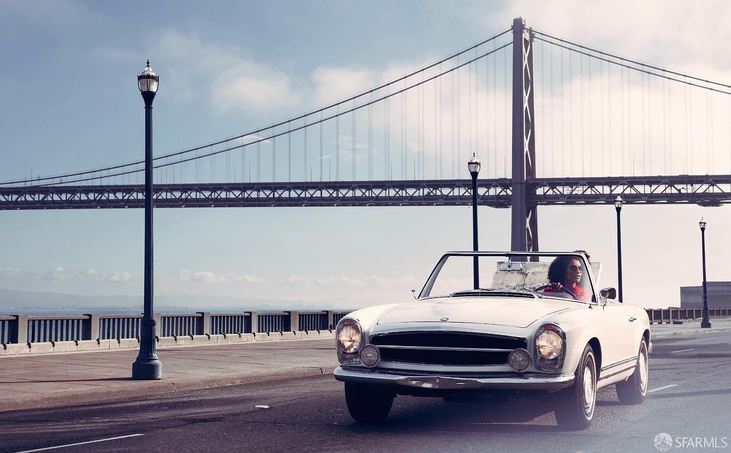 The image showcases a vintage white convertible driving on a road with the Bay Bridge in the background. The car is the main focal point, suggesting a lifestyle of luxury and freedom. The scene is bathed in warm light, creating a nostalgic and aspirational atmosphere.