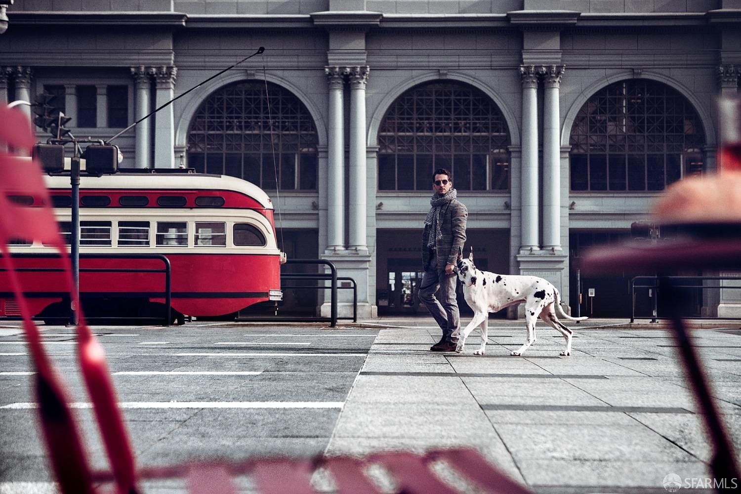 This image showcases the front of a building with prominent architectural features, including arched windows and stately columns. A vintage red and white streetcar is parked nearby, adding a touch of historical charm. A man walks a Great Dane across the crosswalk, creating a sense of urban sophistication and lifestyle appeal.