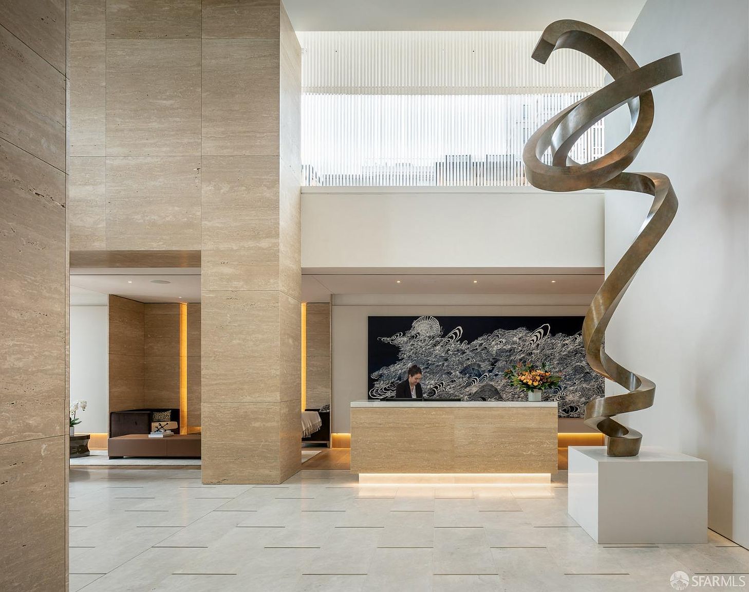 This is an interior shot of a luxurious lobby or hallway, featuring travertine walls and flooring. A modern, abstract sculpture stands prominently near the reception desk, which is illuminated by subtle lighting. The space exudes a sense of sophistication and high-end design, creating a welcoming and impressive atmosphere.