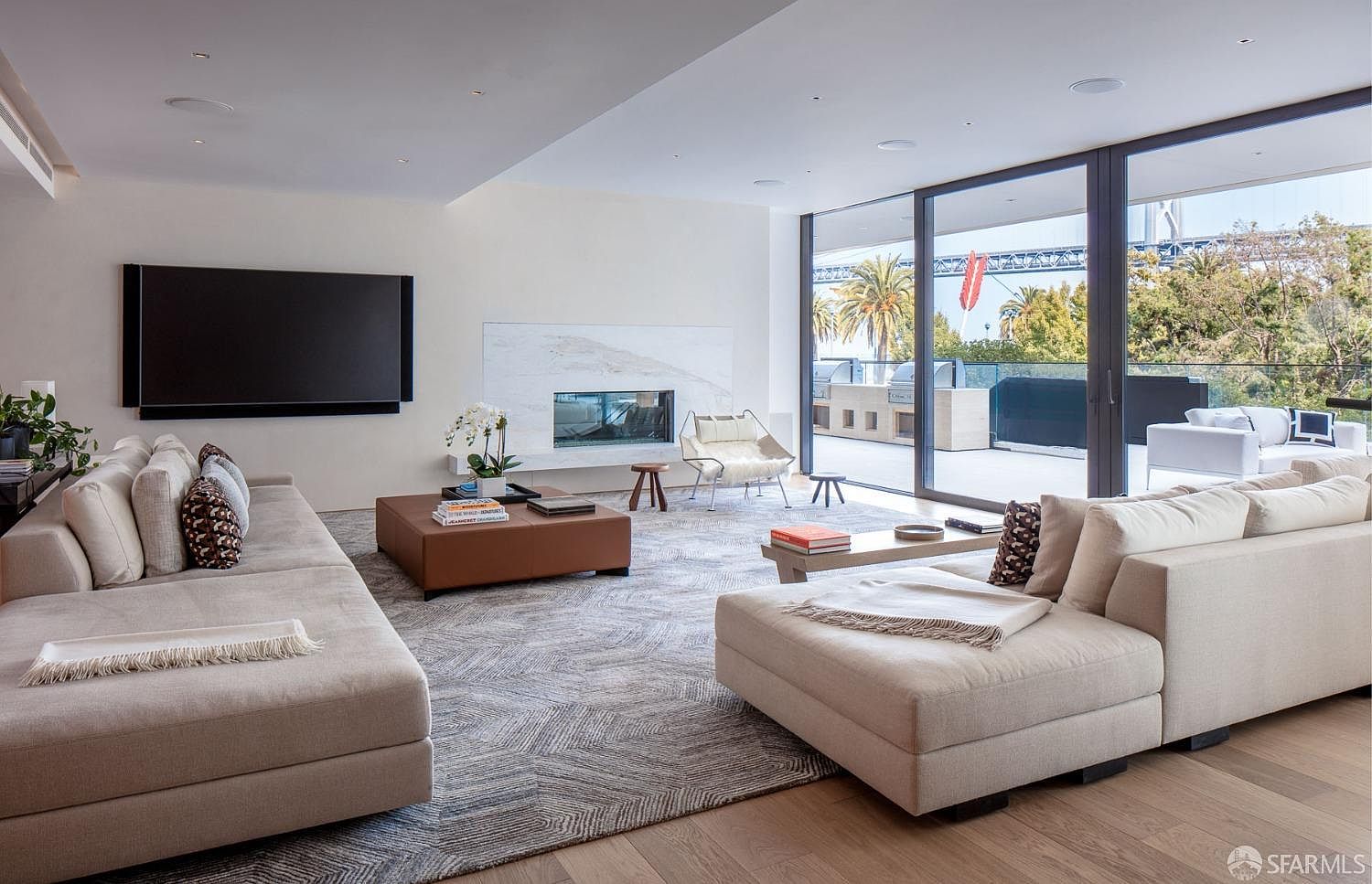 This is a bright and modern living room featuring two large sofas, a fireplace, and floor-to-ceiling windows that offer a view of the outdoors. The room is decorated in neutral tones with a large area rug and hardwood floors. The space is open and airy, creating a comfortable and inviting atmosphere.