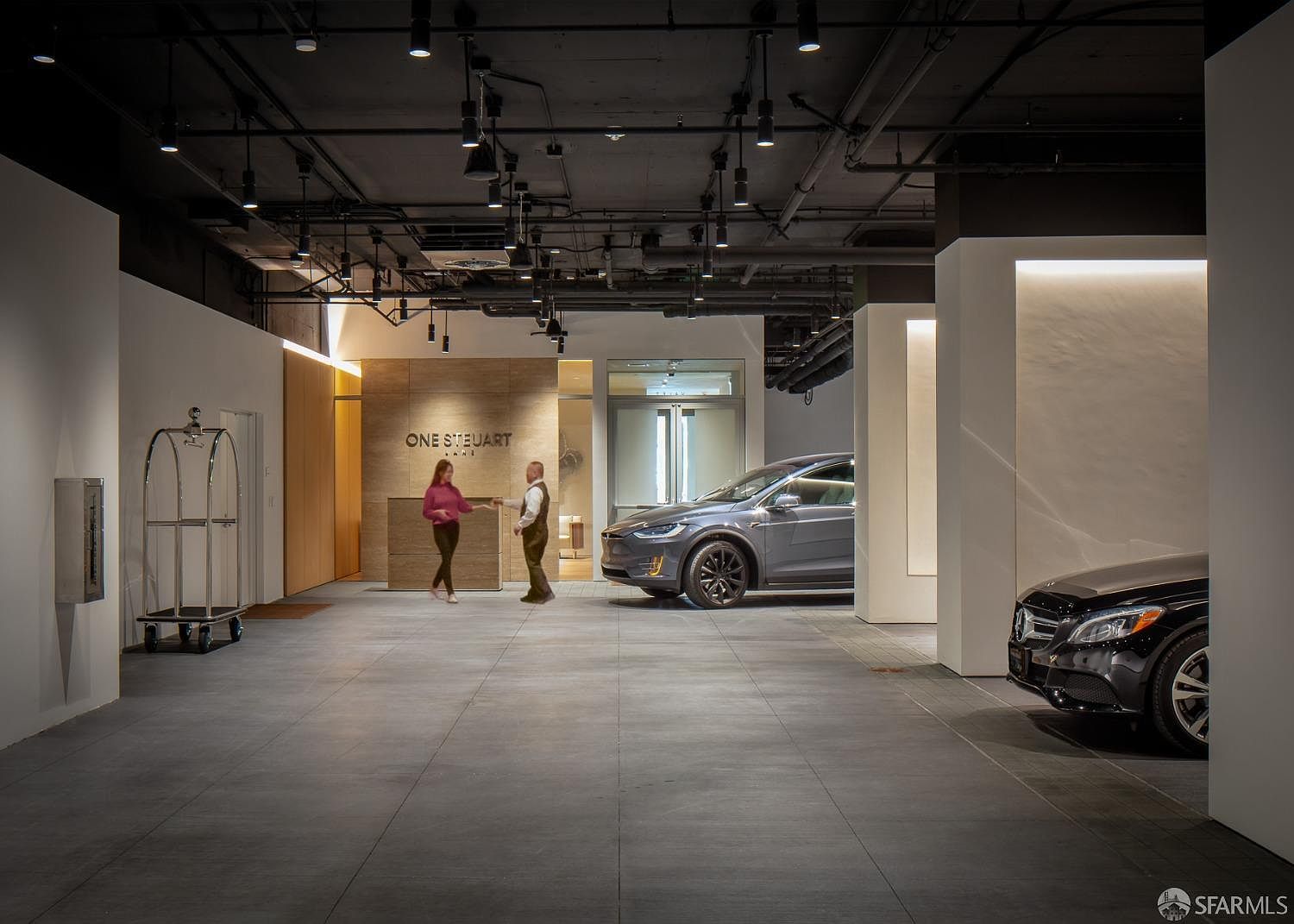 This interior shot showcases a modern lobby or hallway with a sleek, minimalist design. The space features a reception area with the words "ONE STEWART LANE" on the wall, a gray Tesla car, and a black Mercedes car. The lighting is a mix of recessed and track lighting, creating a sophisticated and inviting atmosphere.