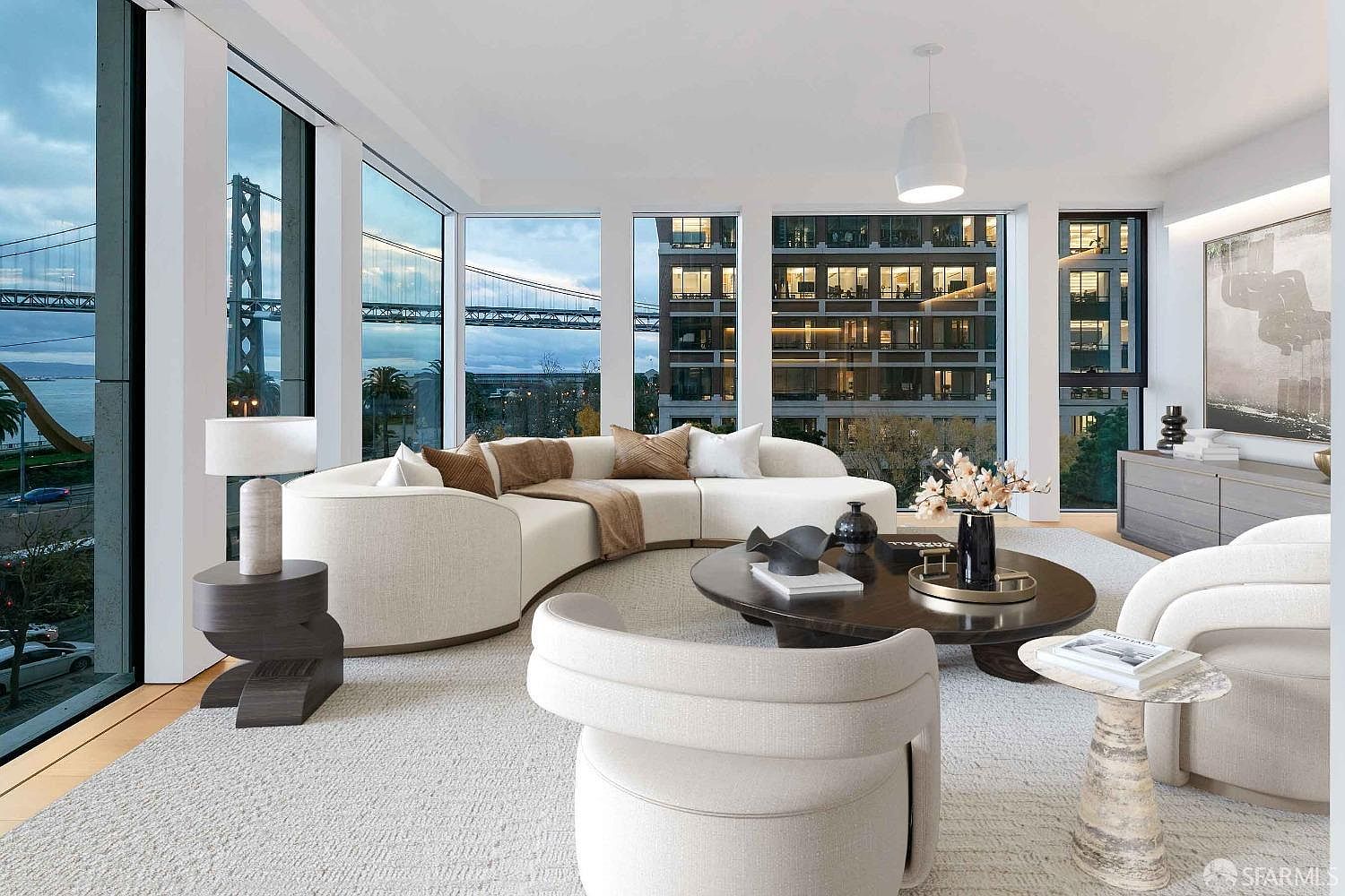 This is a luxurious living room featuring a curved white sofa with decorative pillows, a round dark wood coffee table with decorative objects, and a light-colored rug. Large windows offer stunning views of a bridge and city buildings, enhancing the room's modern and sophisticated ambiance. The room is well-lit and appears spacious, creating an inviting atmosphere.