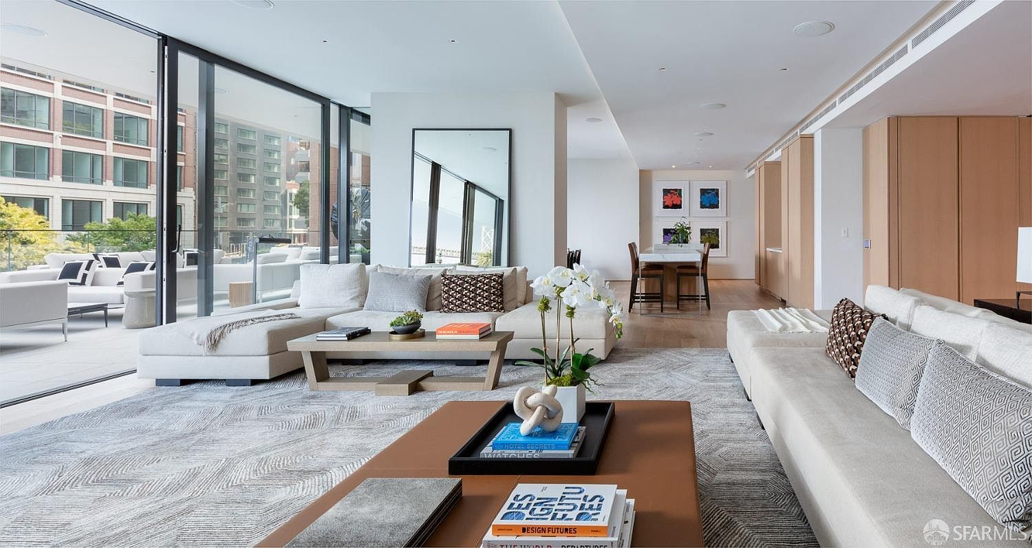 This is a bright and spacious living room featuring modern design elements. The room is furnished with two large sofas, a light-colored area rug, and two coffee tables. Large windows offer views of the exterior, and the open layout connects to a dining area, creating an inviting and luxurious atmosphere.