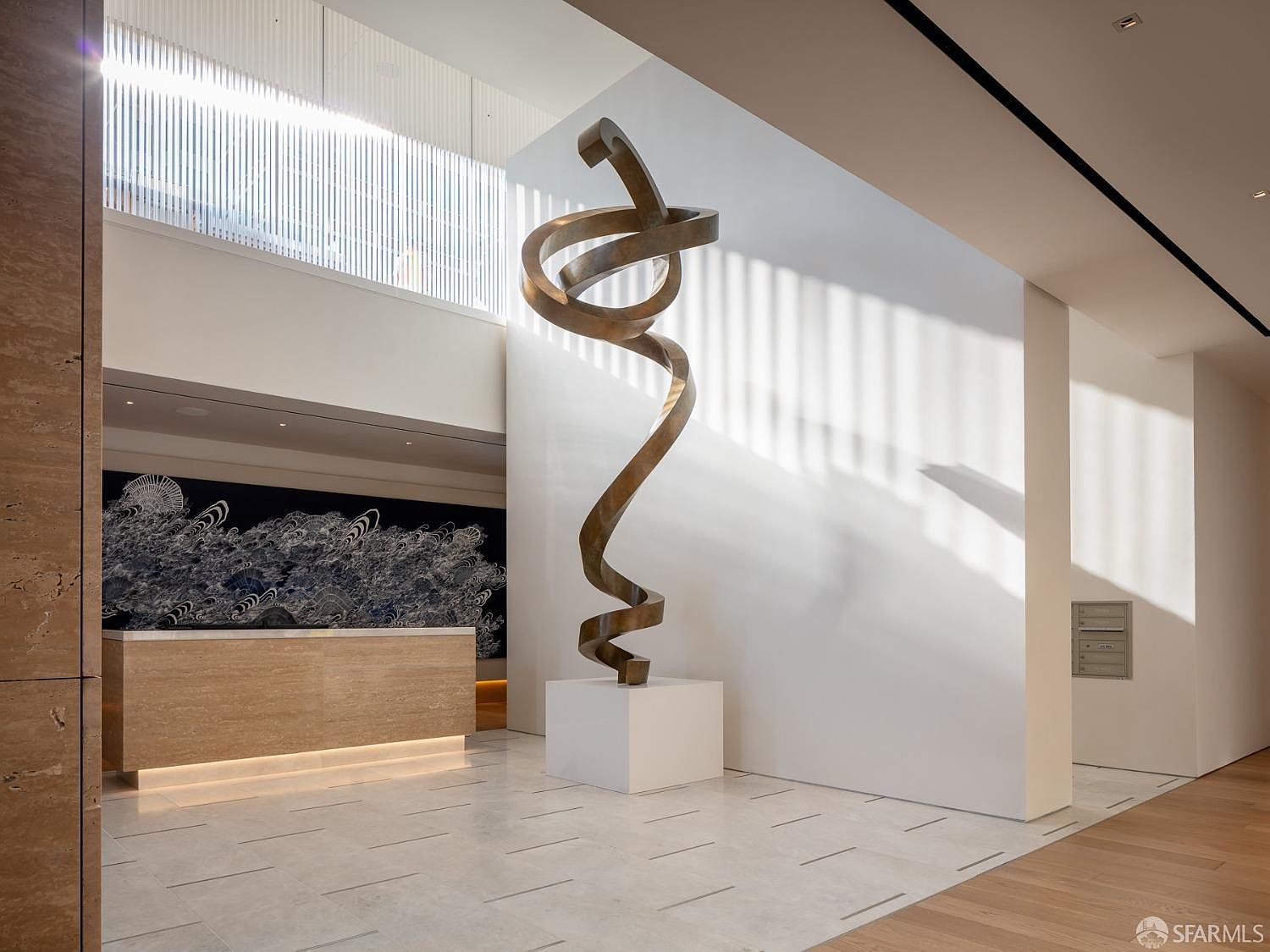 This interior shot showcases a modern hallway or lobby area with a striking bronze sculpture as a focal point. The space features clean lines, a neutral color palette, and a combination of tile and wood flooring. Natural light streams in through a high window, creating a bright and airy atmosphere, while a reception desk is visible in the background.