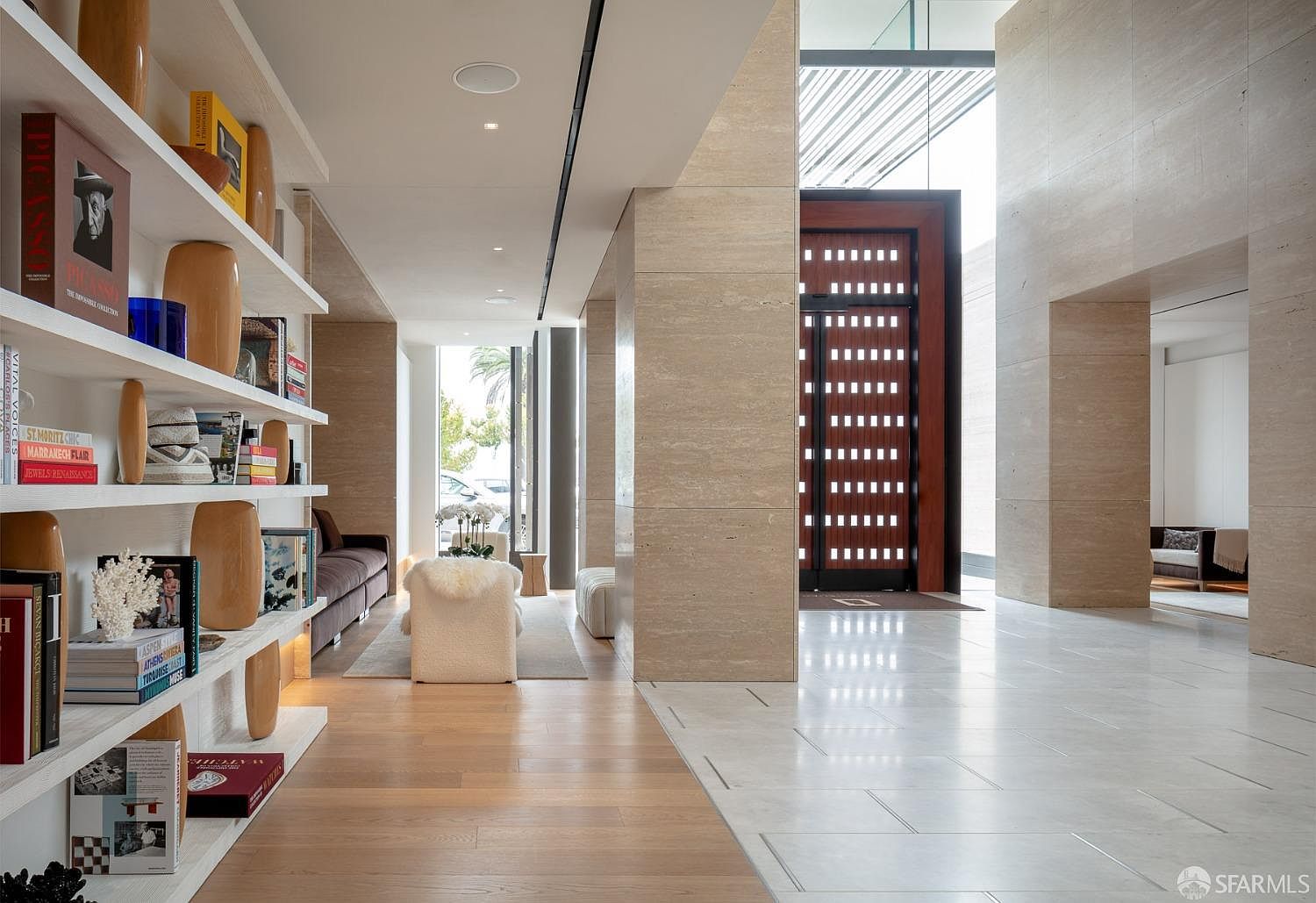 This interior shot showcases a luxurious hallway with a blend of wood and tile flooring, leading to a bright entryway. The space is defined by travertine-clad walls and columns, complemented by built-in shelving displaying books and decorative items. A modern, minimalist aesthetic is evident throughout, emphasizing clean lines and natural light.