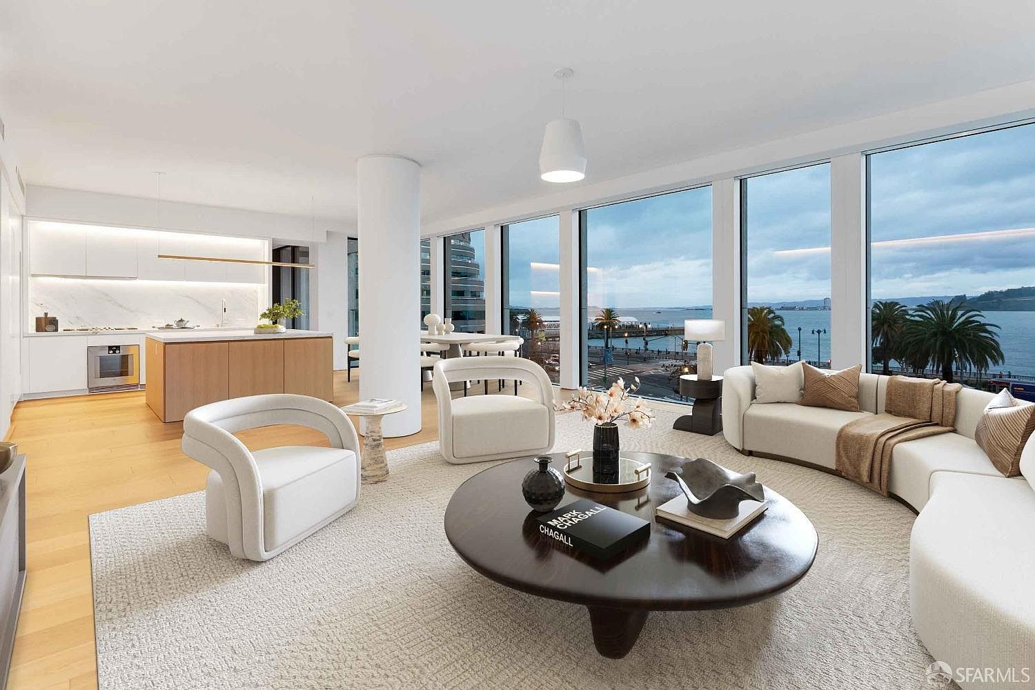 This is a bright and airy living room with floor-to-ceiling windows offering stunning waterfront views. The room features modern, curved furniture including a white sofa and armchairs, complemented by a dark wood round coffee table. The open-concept design seamlessly integrates the living area with a modern kitchen, creating a sophisticated and inviting space.