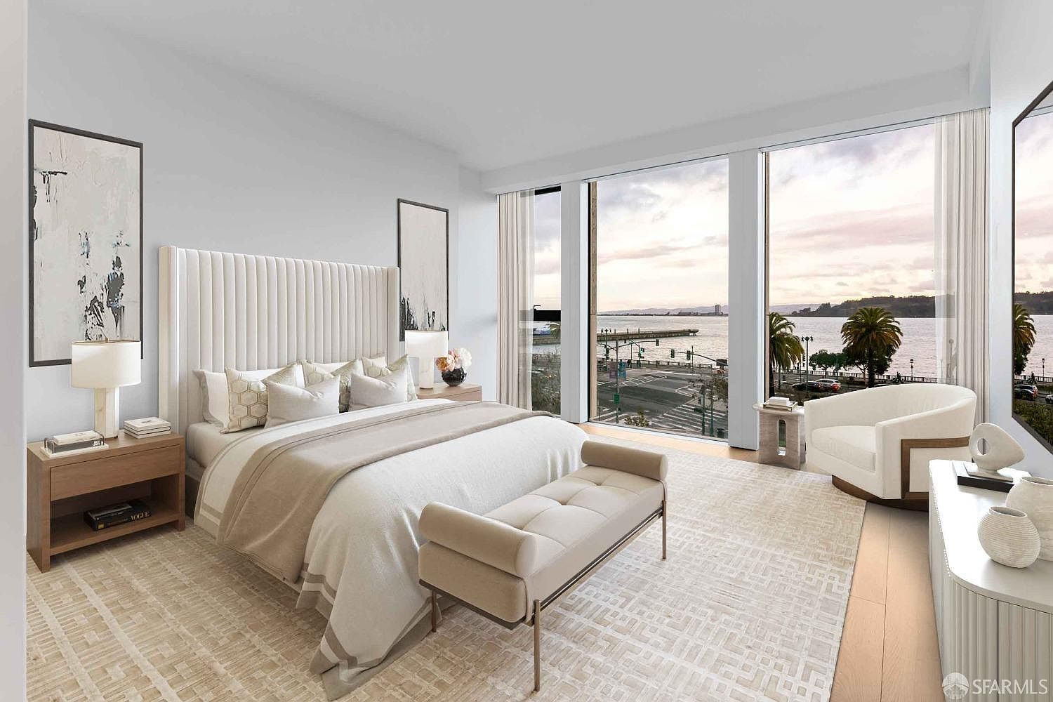 This is a bright and airy primary bedroom featuring a tall, upholstered headboard and a neutral color palette. Large windows offer a stunning view of the waterfront, enhancing the room's appeal. A bench sits at the foot of the bed, and the room is decorated with modern art and stylish furniture, creating a serene and luxurious atmosphere.